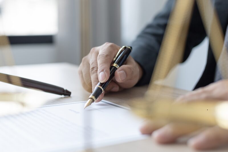 Dettaglio di un braccio con cravatta che scrive e un contratto con penna stilosa.
