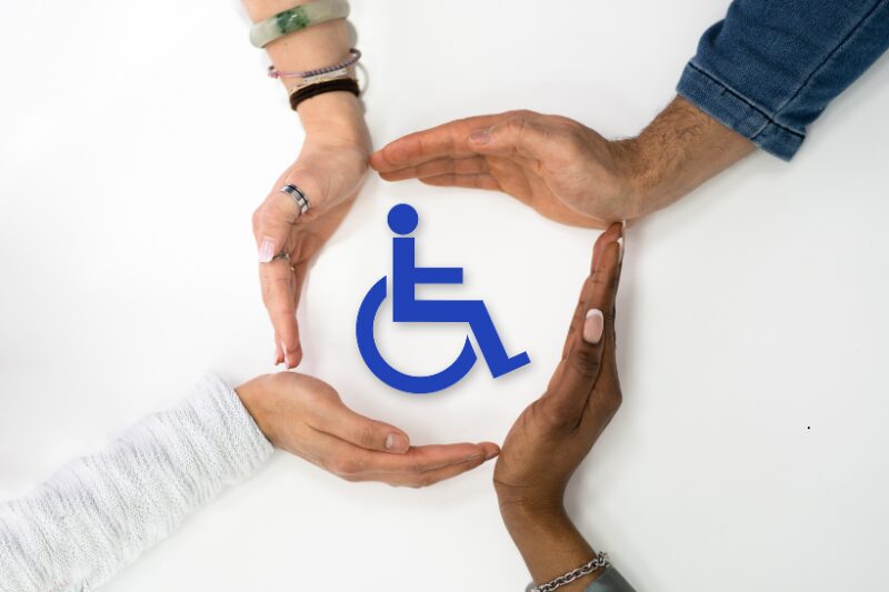 Quattro mani di persone diverse per età e carnagione formano un cerchio protettivo attorno al simbolo della disabilità, esprimendo inclusione, sostegno e solidarietà.