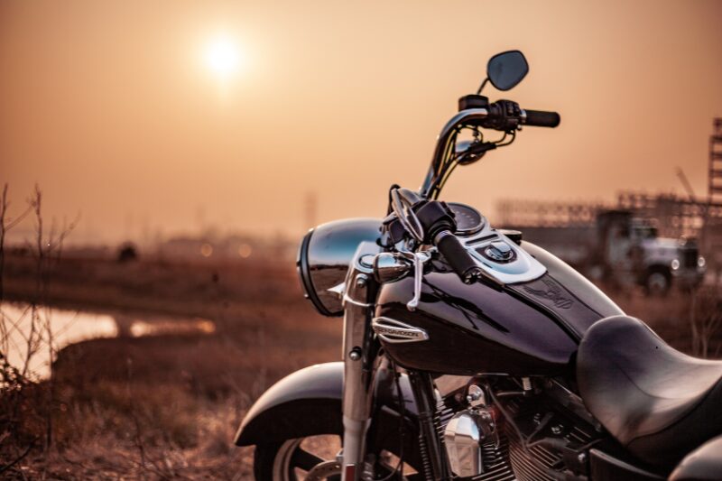 Moto custom parcheggiata su un terreno all’aperto al tramonto, con il sole basso sull’orizzonte e una luce calda che avvolge il paesaggio rurale sullo sfondo.