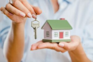 Primo piano di una persona che tiene in una mano una piccola casa giocattolo e nell'altra una chiave, simbolo della consegna o dell’acquisto di un’abitazione.
