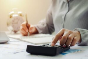 Persona che calcola delle spese o bilanci con una calcolatrice su una scrivania, prendendo appunti su un blocco note. Sullo sfondo si intravedono un orologio e grafici con dati.