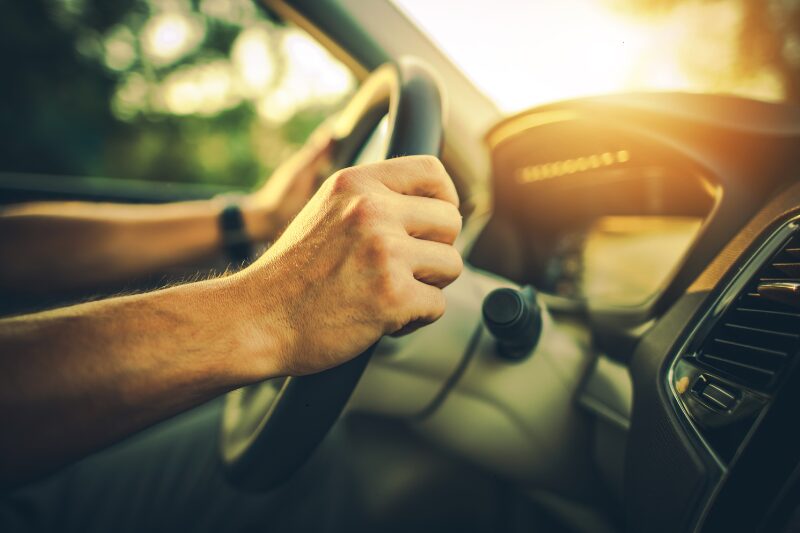 Dettaglio delle mani di un neopatentato sul volante di un’auto, illuminate dalla luce calda del tramonto. Interni dell’auto nuovi