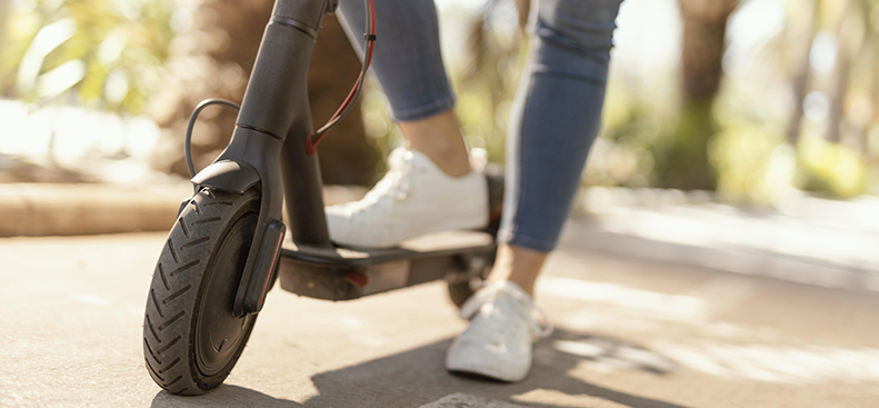 Persona guida un monopattino elettrico su una pista ciclabile in città, indossando scarpe da ginnastica bianche e jeans. L’immagine rappresenta la mobilità urbana sostenibile e l’uso dei monopattini elettrici come mezzo di trasporto ecologico.