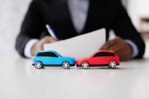 Due modellini di auto, una blu e una rossa, che simulano un incidente su un tavolo con una persona sullo sfondo che compila un modulo assicurativo.