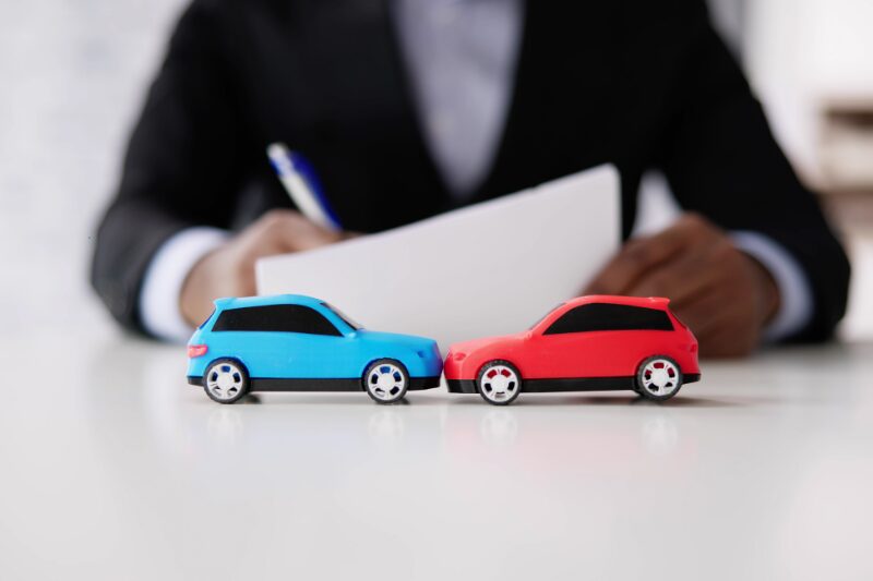 Due modellini di auto, una blu e una rossa, che simulano un incidente su un tavolo con una persona sullo sfondo che compila un modulo assicurativo.