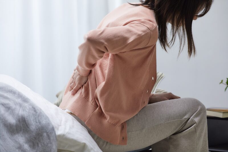 Donna seduta sul letto che si tiene la schiena dolorante, indicando un possibile infortunio domestico.