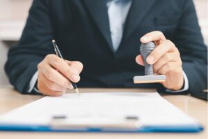 Una persona in giacca sta firmando un documento su una scrivania, tenendo in mano una penna e un timbro.
