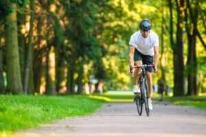 Ciclista adulto con casco che pedala su una pista ciclabile immersa nel verde in un parco urbano.