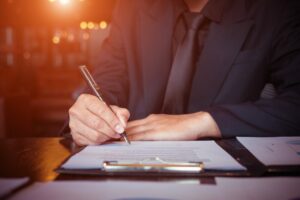 Uomo in giacca e cravatta che firma un documento assicurativo su una scrivania con una penna.