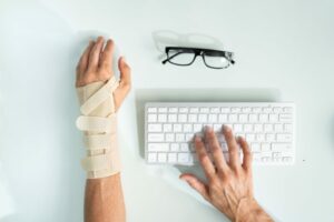 Persona al computer con un tutore ortopedico al polso mentre digita sulla tastiera, con occhiali appoggiati accanto.