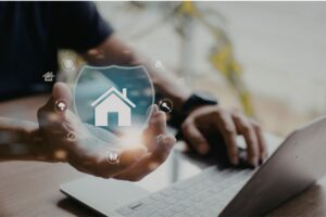 Mano che protegge un’icona di casa all’interno di uno scudo digitale mentre una persona lavora al computer, concetto di assicurazione e tutela dei beni.