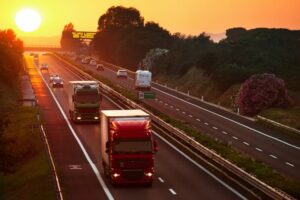 Camion e automobili in viaggio su un’autostrada al tramonto, con il sole basso all’orizzonte e traffico scorrevole in entrambe le direzioni.