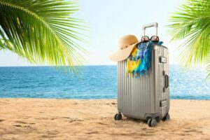 Valigia sulla spiaggia con cappello e accessori da vacanza davanti al mare, immagine che rappresenta viaggi e soggiorni sereni protetti da una copertura assicurativa.