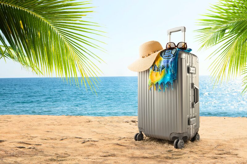 Valigia sulla spiaggia con cappello e accessori da vacanza davanti al mare, immagine che rappresenta viaggi e soggiorni sereni protetti da una copertura assicurativa.