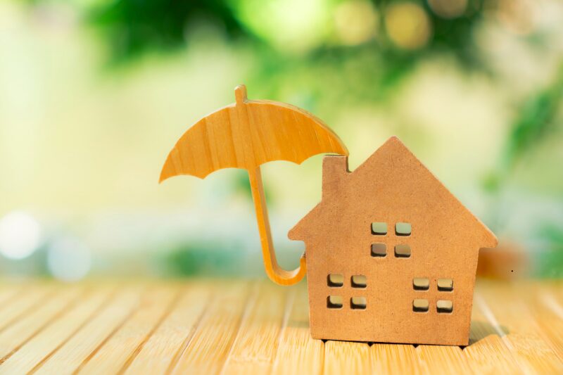 Casetta in legno protetta da un ombrello, simbolo di assicurazione casa contro eventi atmosferici e danni climatici.