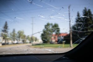 Una veduta dall'interno dell'abitacolo di un'auto attraverso un parabrezza pesantemente rotto con un motivo di crepe a ragnatela, con alberi, una strada e pali della luce visibili all'esterno.