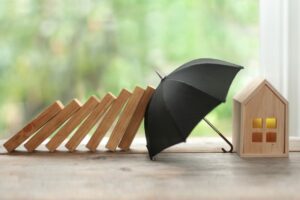 Una casetta in legno protetta da un piccolo ombrello nero che blocca una fila di blocchi di legno mentre cadono a domino, su uno sfondo luminoso con piante.